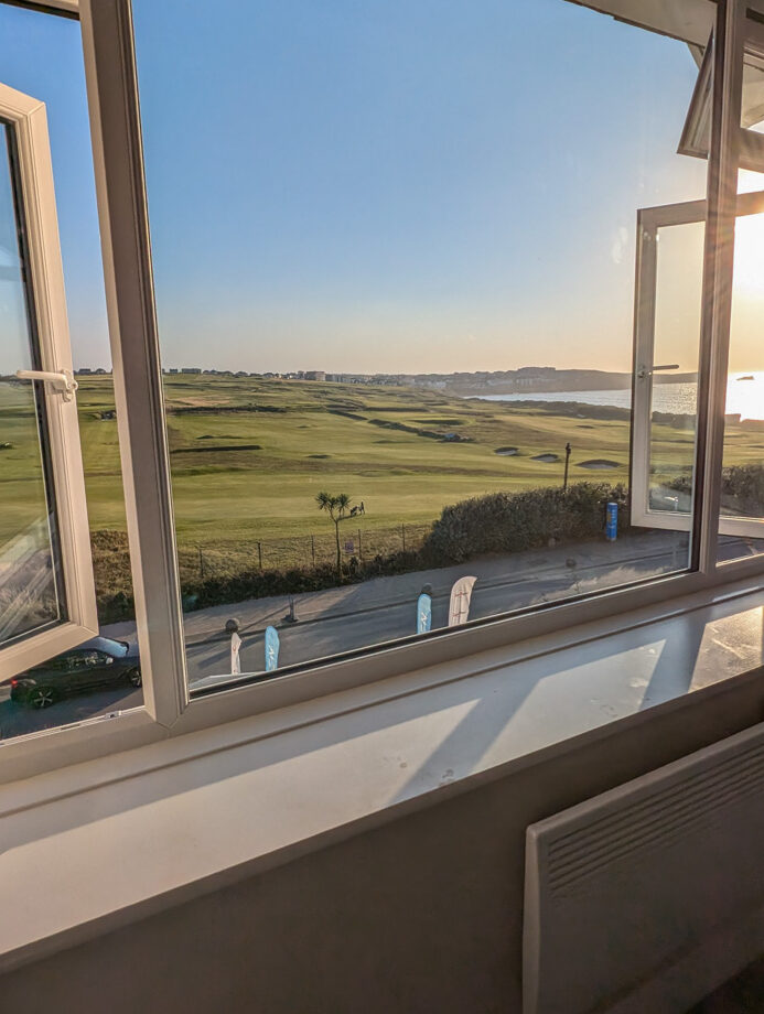 A wide-open window frames a view of a green golf course stretching towards a calm blue ocean, under a clear sky with sunlight glinting in from the side.