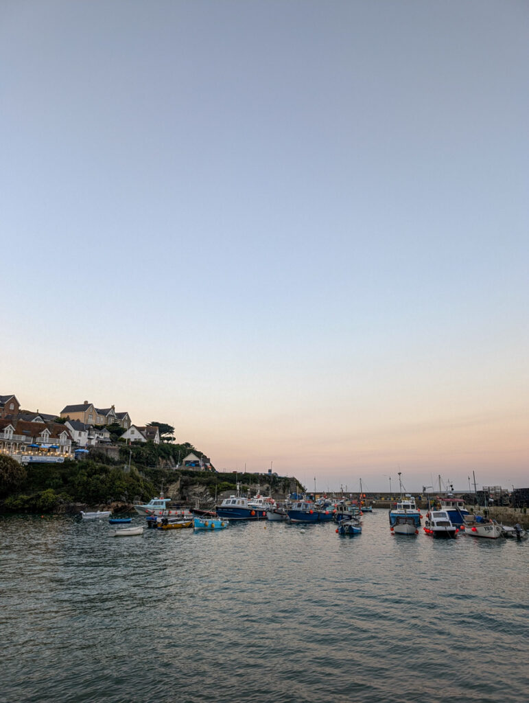 A peaceful harbor scene at dusk, with several colorful boats anchored in calm waters and charming hillside houses in the background against a soft, pastel sky.