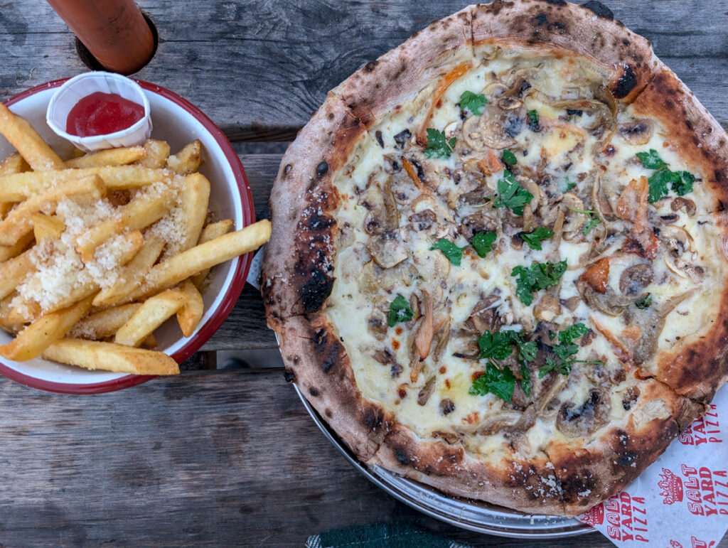 A close-up of a rustic wood-fired pizza topped with mushrooms and herbs alongside a bowl of golden fries sprinkled with parmesan and served with a side of ketchup.