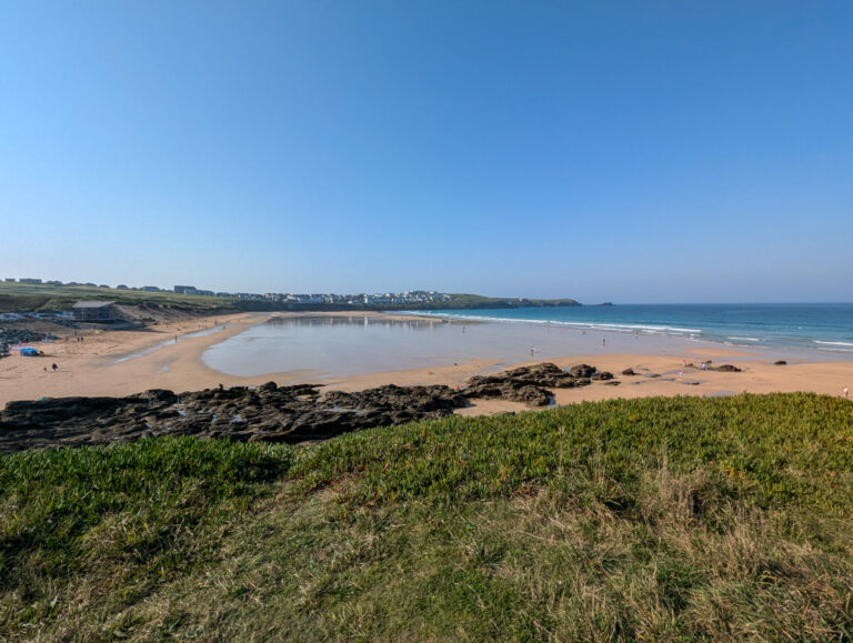 A wide-angle view of Fistral Beach showing sandy shores, gentle waves, and a few beachgoers enjoying the sunny weather, with grassy dunes and a distant townscape.