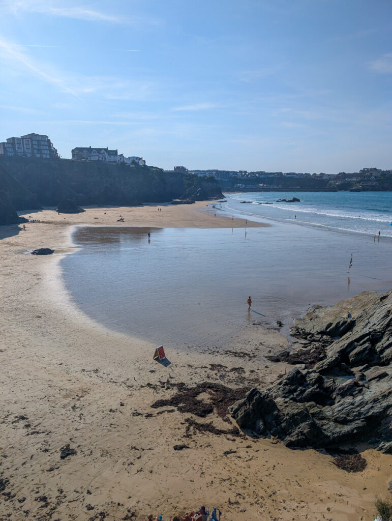 A serene cove with golden sand bordered by rocky cliffs, with a few people walking along the shore under a bright blue sky.