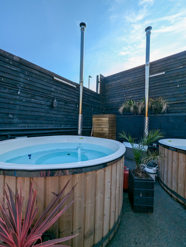 A rustic outdoor setting with a round, wood-panel hot tub under a clear sky, accompanied by a wood storage area and cozy seating.