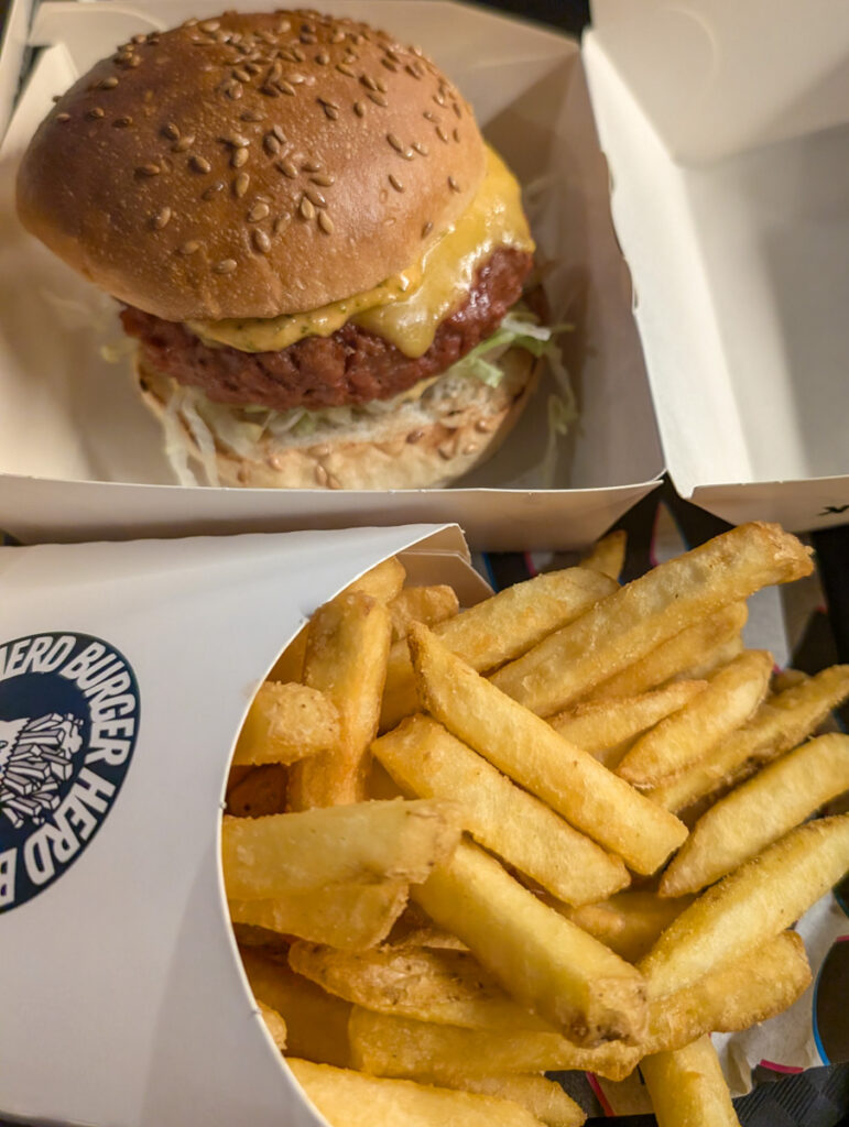 A juicy burger with melted cheese in a takeout box, accompanied by a side of thick-cut fries in a branded cup, ready to enjoy.